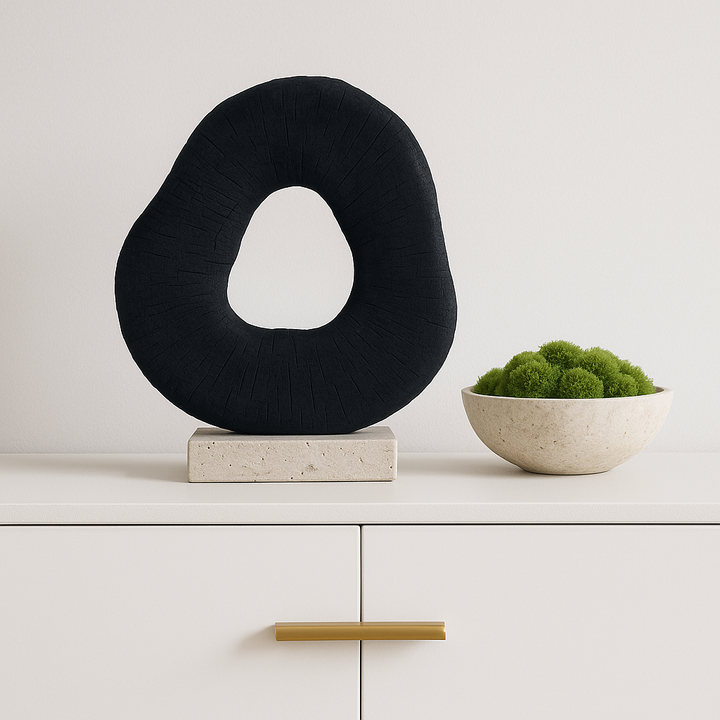Square luxury lifestyle image of black organic ring sculpture on travertine base beside a stone bowl filled with soft green moss on a white sideboard with gold handle.