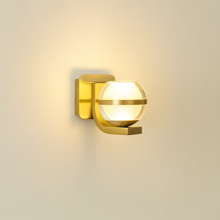 Modern brass wall light with clear glass globe casting a warm glow on a neutral wall