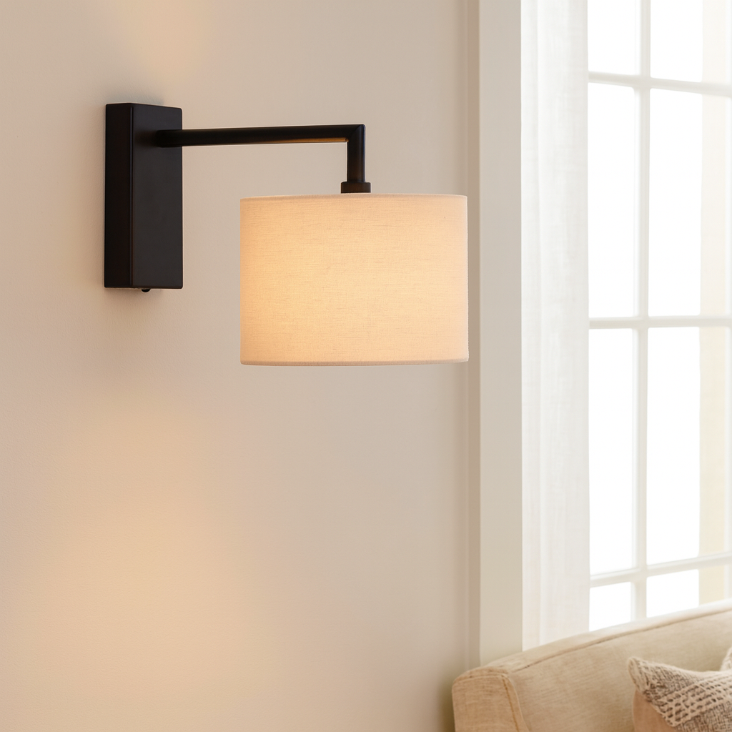 Luxury lifestyle image of a Fairview black wall light with a 25cm white fabric shade mounted on a neutral wall beside a window and sofa.