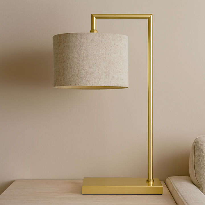 Lifestyle image of the Fairview gold table lamp with a 25cm natural fabric shade and 59cm total height, styled on a light wooden surface beside a neutral sofa.