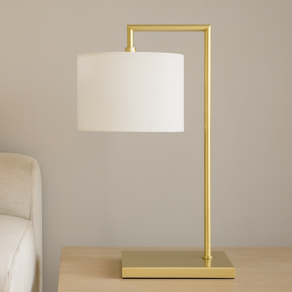 Lifestyle image of the Fairview gold table lamp with a 25cm white fabric shade and 59cm total height, styled on a wooden side table beside a neutral sofa.