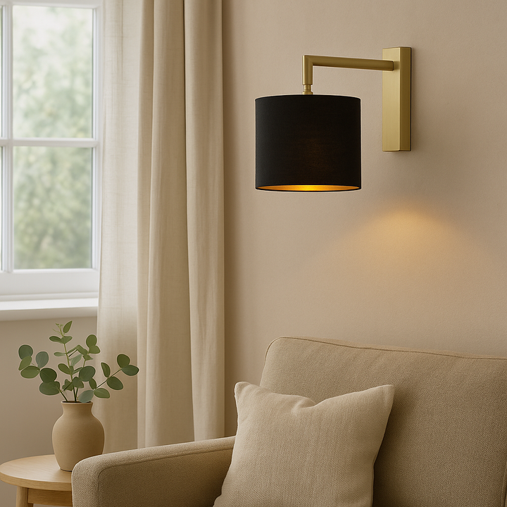 Lifestyle image of the Fairview gold wall light with a 25cm black fabric shade and gold lining, mounted on a neutral wall beside a beige sofa and window with curtains.