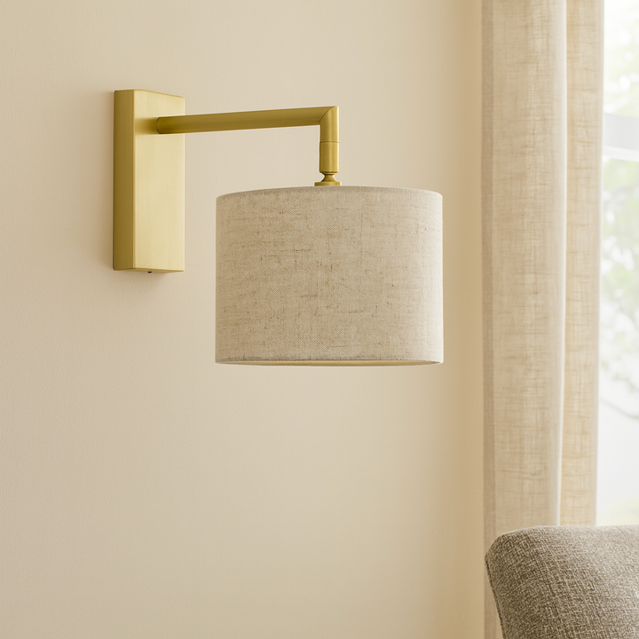 Luxury lifestyle image of the Fairview gold wall light with a 25cm natural fabric drum shade mounted on a neutral wall beside a window and armchair