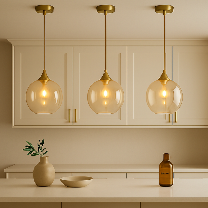 Square lifestyle image of three Irvine gold amber glass pendant lights above a cream kitchen island with gold cabinet accents and minimal decorative styling.