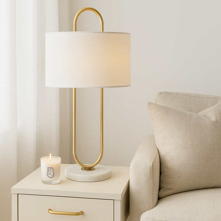 Modern gold table lamp with white shade on cream side table with gold handle, styled with a lit designer candle in a bright living room.