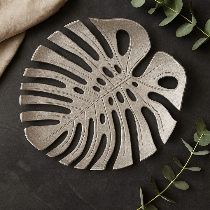 Overhead view of luxury silver monstera leaf decorative dish styled on dark stone surface with fabric and eucalyptus leaves for modern dΓ©cor