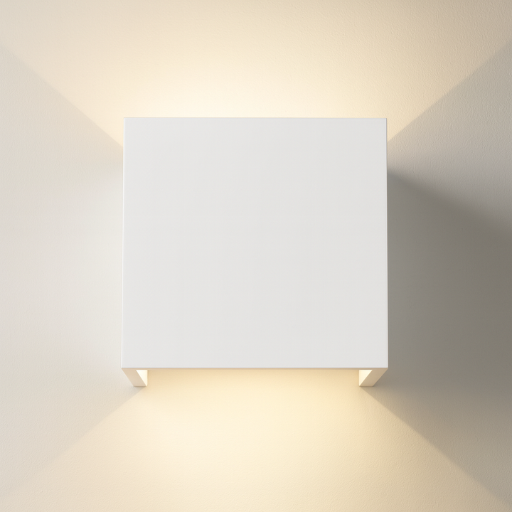 Square ceramic wall light in a smooth white finish, mounted on a white wall with warm up and down lighting creating soft triangular patterns.