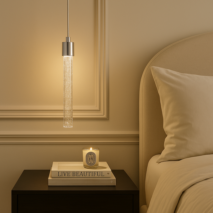 Luxury bedroom scene featuring the Sydney Single Pendant in polished silver, styled as a bedside light above a dark wood table with stacked books and a lit candle, set against cream wall panelling and neutral bedding.