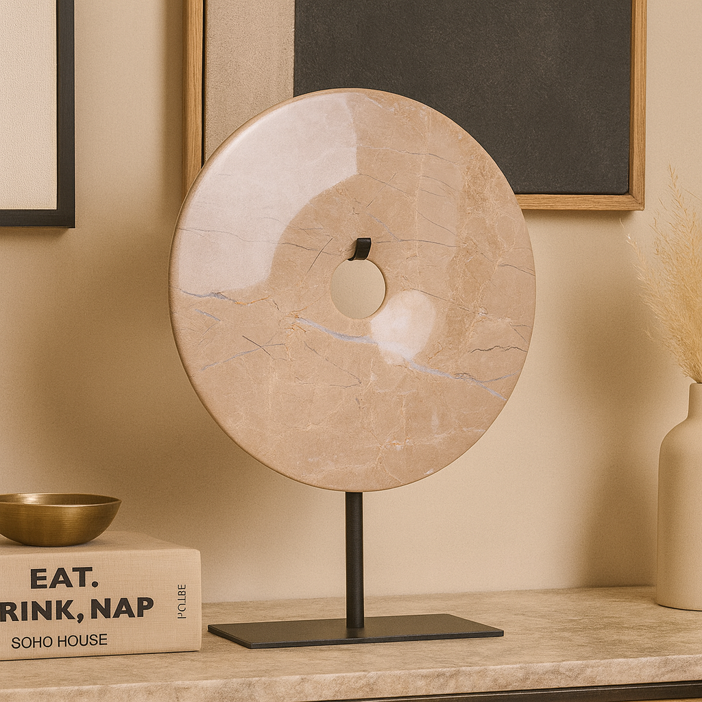 Beige marble disc sculpture with natural veining on black stand, styled on a console table with books, brass bowl, and pampas grass in a Scandi-luxury interior