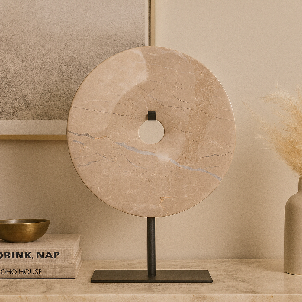 Beige marble disc sculpture with natural veining on a black stand, styled on a console table with books, brass bowl, and pampas grass in a Scandi-luxury setting