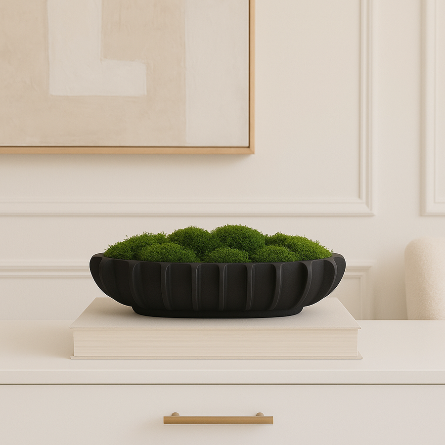 Black fluted bowl filled with green moss on a large book and white console table.