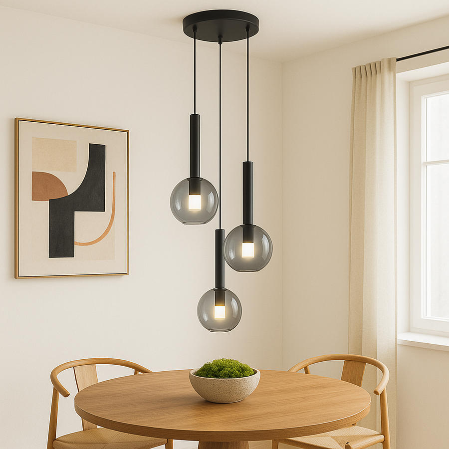 Modern black and glass pendant light with three globes hanging above a round wooden dining table with green moss bowl, light wood chairs, abstract artwork, and light walls in a bright dining room.