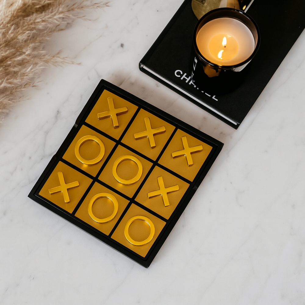Flat lay of luxury gold noughts and crosses game on white marble table with black candle on Chanel book and pampas styling