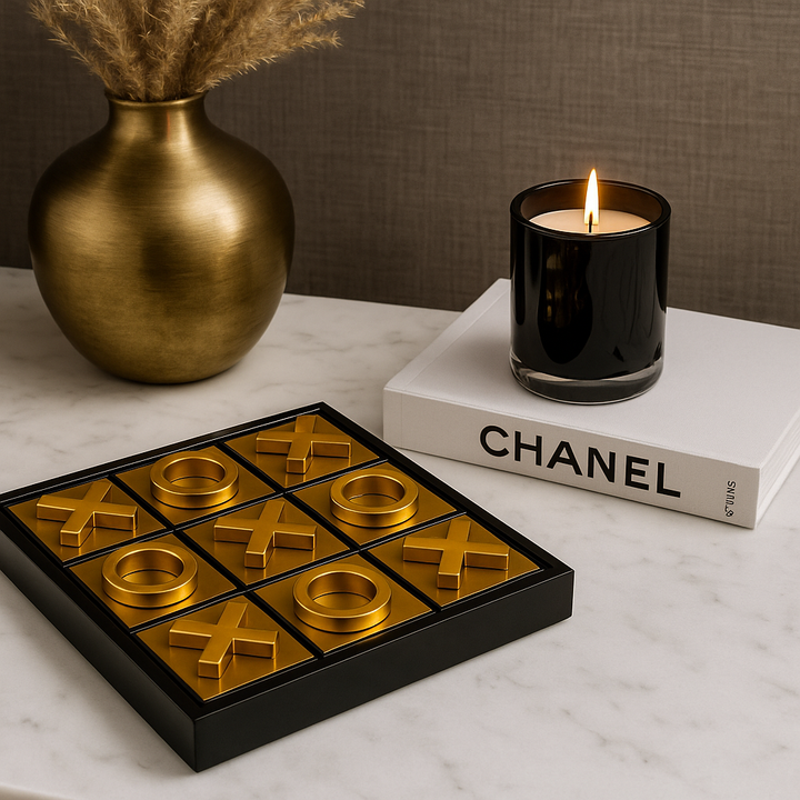 Luxury gold noughts and crosses game on white marble table with black glass candle on Chanel book and gold vase with pampas