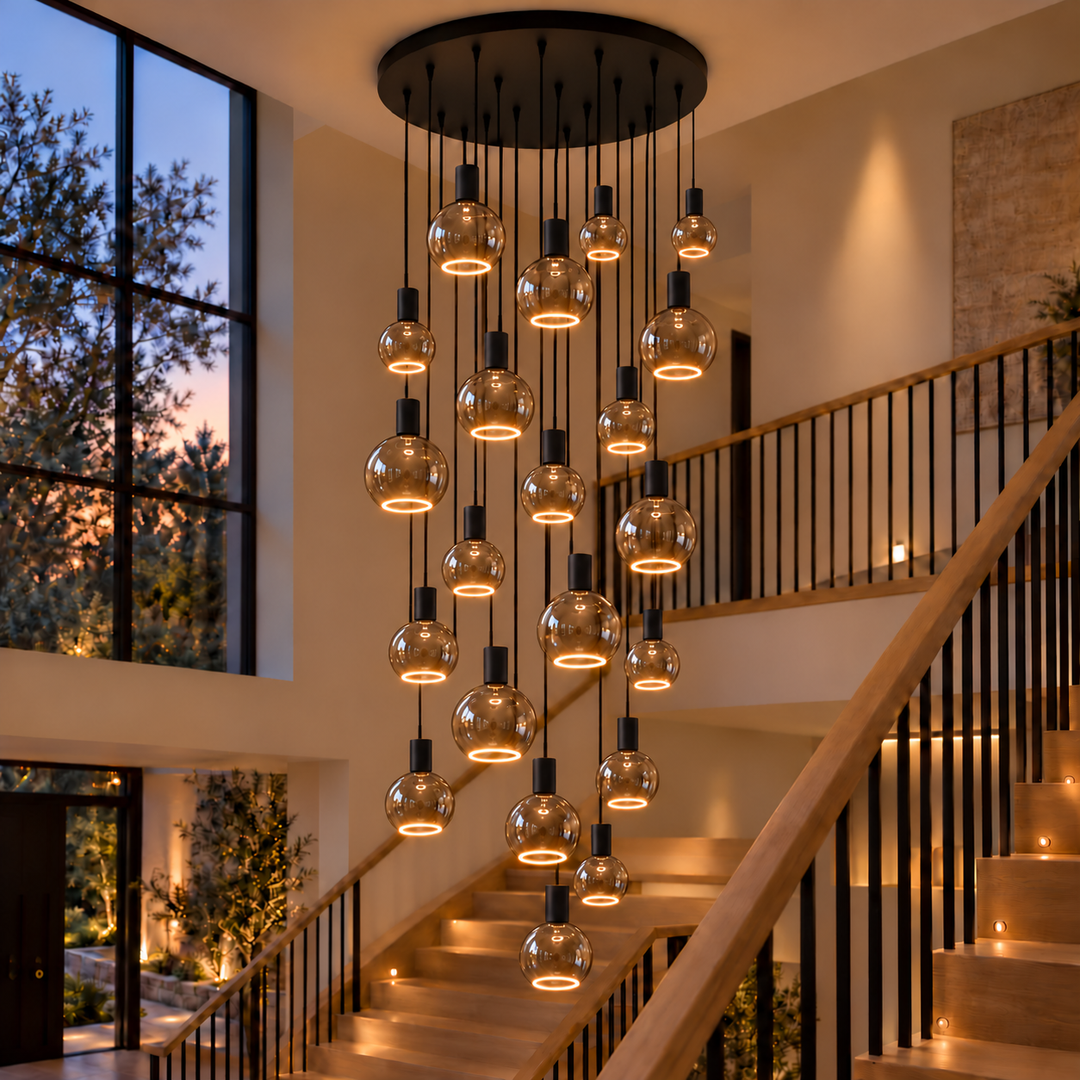 Roma 21-light pendant with black caps and smoked glass bulbs in three sizes, illuminated by warm bottom halo rings above an oak staircase.