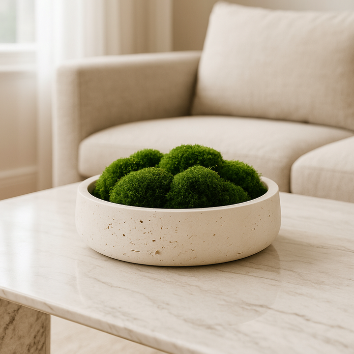 Luxury travertine stone bowl filled with green moss styled on a high-end white marble coffee table in a modern neutral living room.