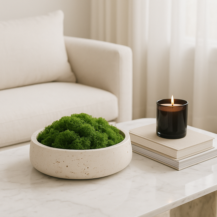 Luxury travertine stone bowl filled with soft green moss on a white marble coffee table, styled with stacked books and a lit dark glass candle in a modern neutral living room.