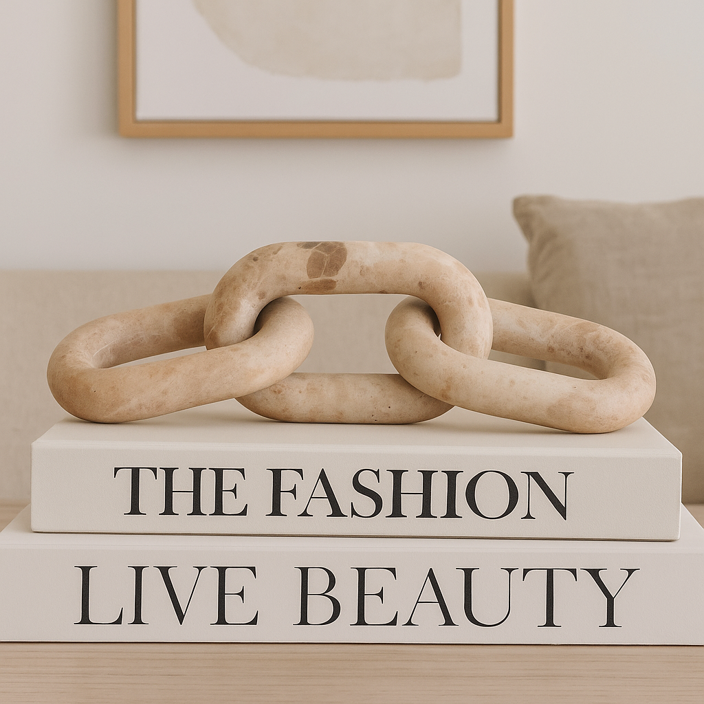 Beige travertine chain link sculpture styled on large cream coffee table books in a modern neutral living room