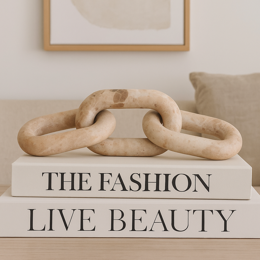 Beige travertine chain link sculpture styled on large cream coffee table books in a modern neutral living room