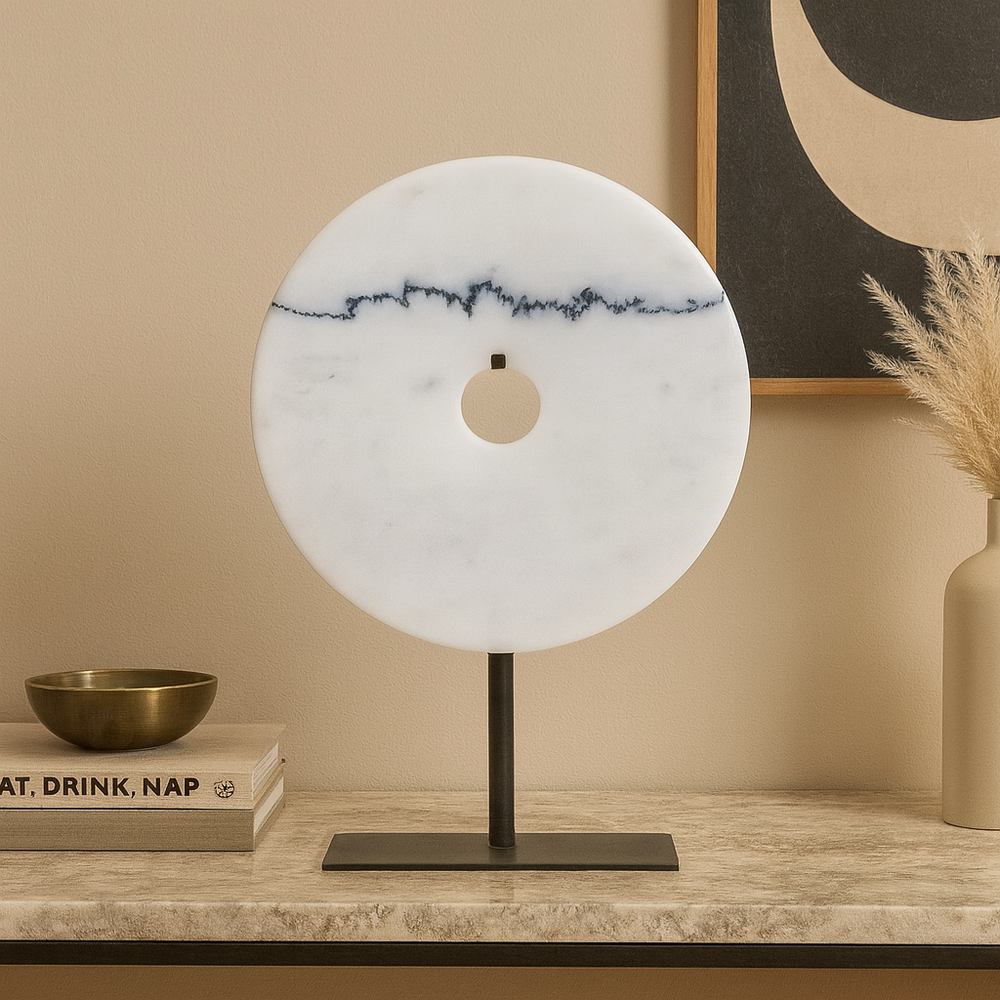 White marble disc sculpture with dark veining detail on a black stand, styled on a luxury console table with books, brass bowl, and vase in a Scandi-luxury setting