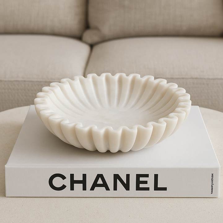 Luxury lifestyle image of a white marble scalloped bowl styled on top of a large Chanel coffee table book in a modern neutral living room