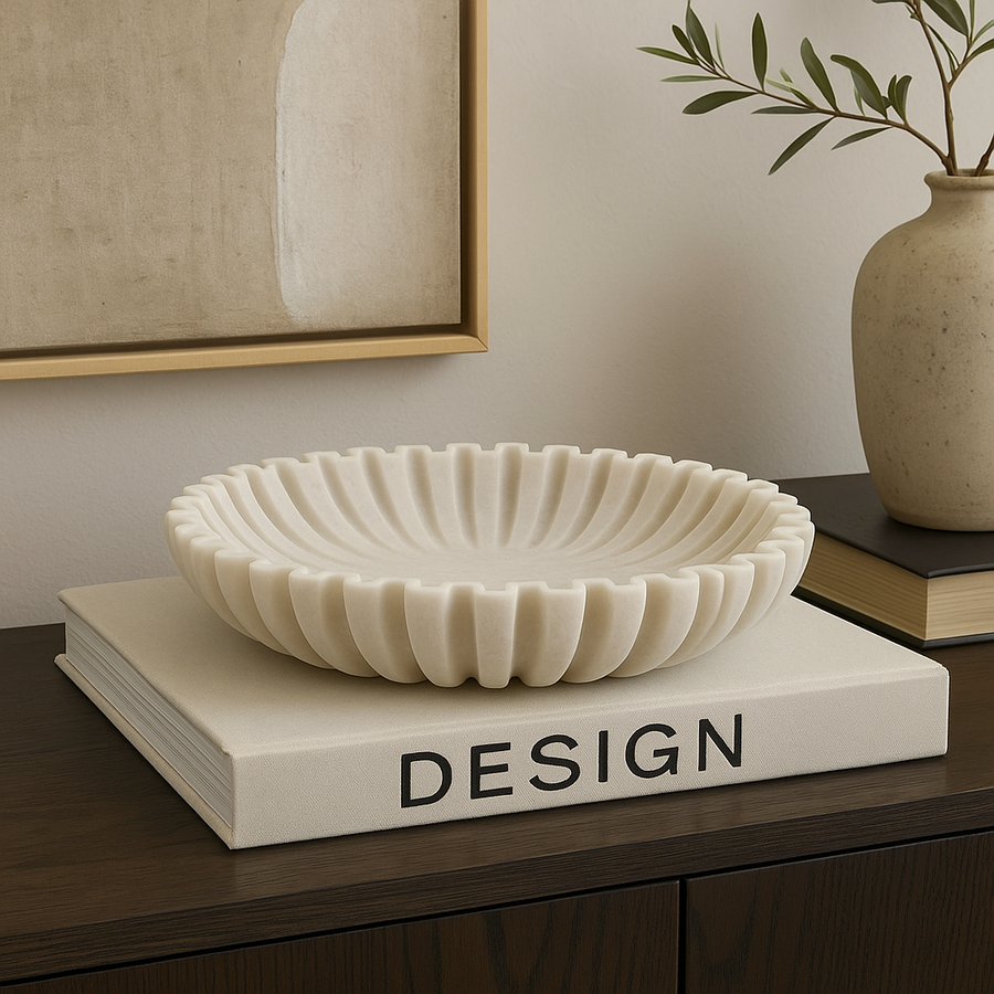 Luxury lifestyle image of a white marble scalloped bowl with no filling, styled on a large design book atop a dark wooden sideboard with artwork and a ceramic vase.
