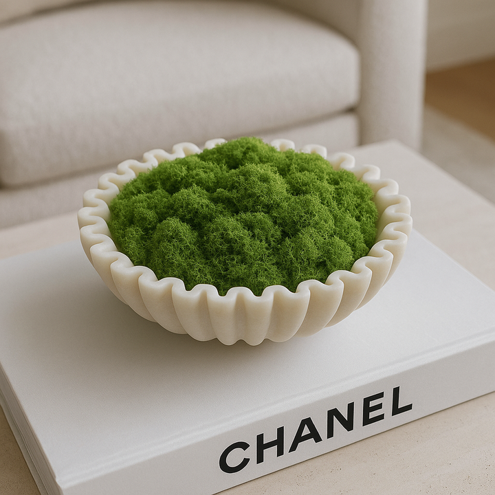 Luxury lifestyle image of a white marble scalloped bowl filled with soft green moss, styled on top of a large Chanel coffee table book in a modern neutral living room
