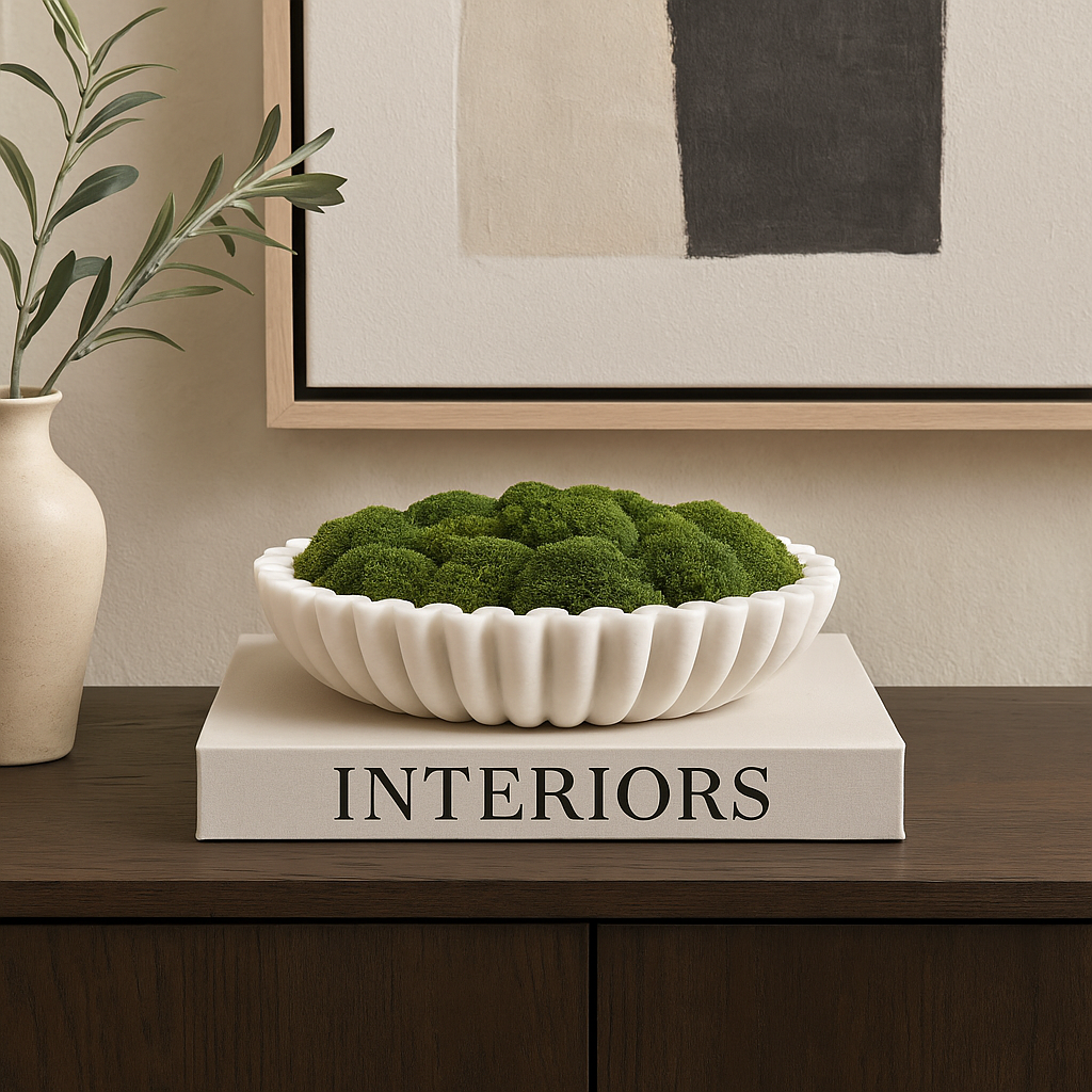 Luxury lifestyle image of a white marble scalloped bowl filled with green moss, styled on a large design book atop a dark wooden sideboard with artwork and a ceramic vase.